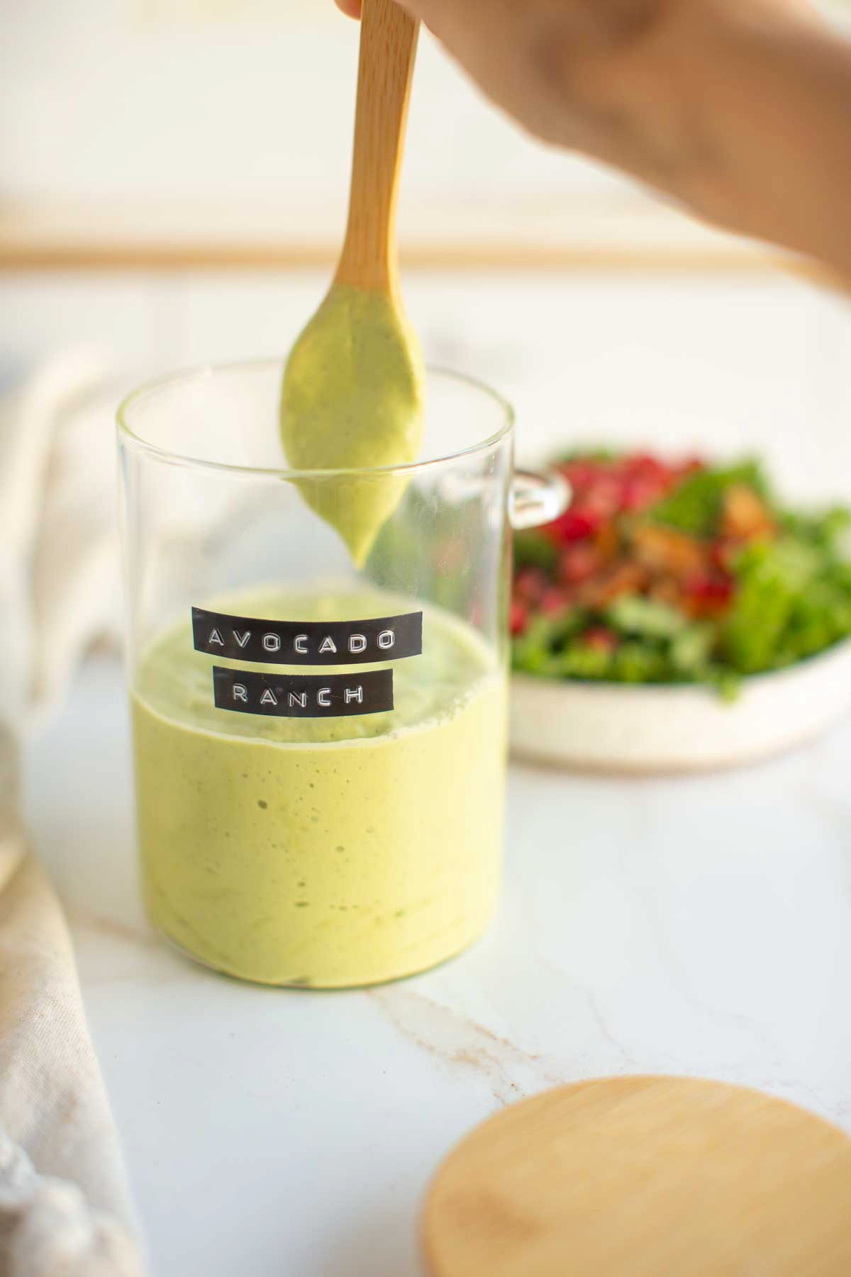 A hand holding a wooden spoon into a glass jar full of fresh avocado ranch dressing and next to a plate full of fresh salad.