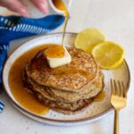 A plate of lemon poppyseed pancakes with a pat of vegan butter, lemon slices, and maple syrup pouring onto the top.