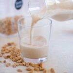 Peanut butter milk pouring from a glass bottle into a small glass with peanuts strewn around it.
