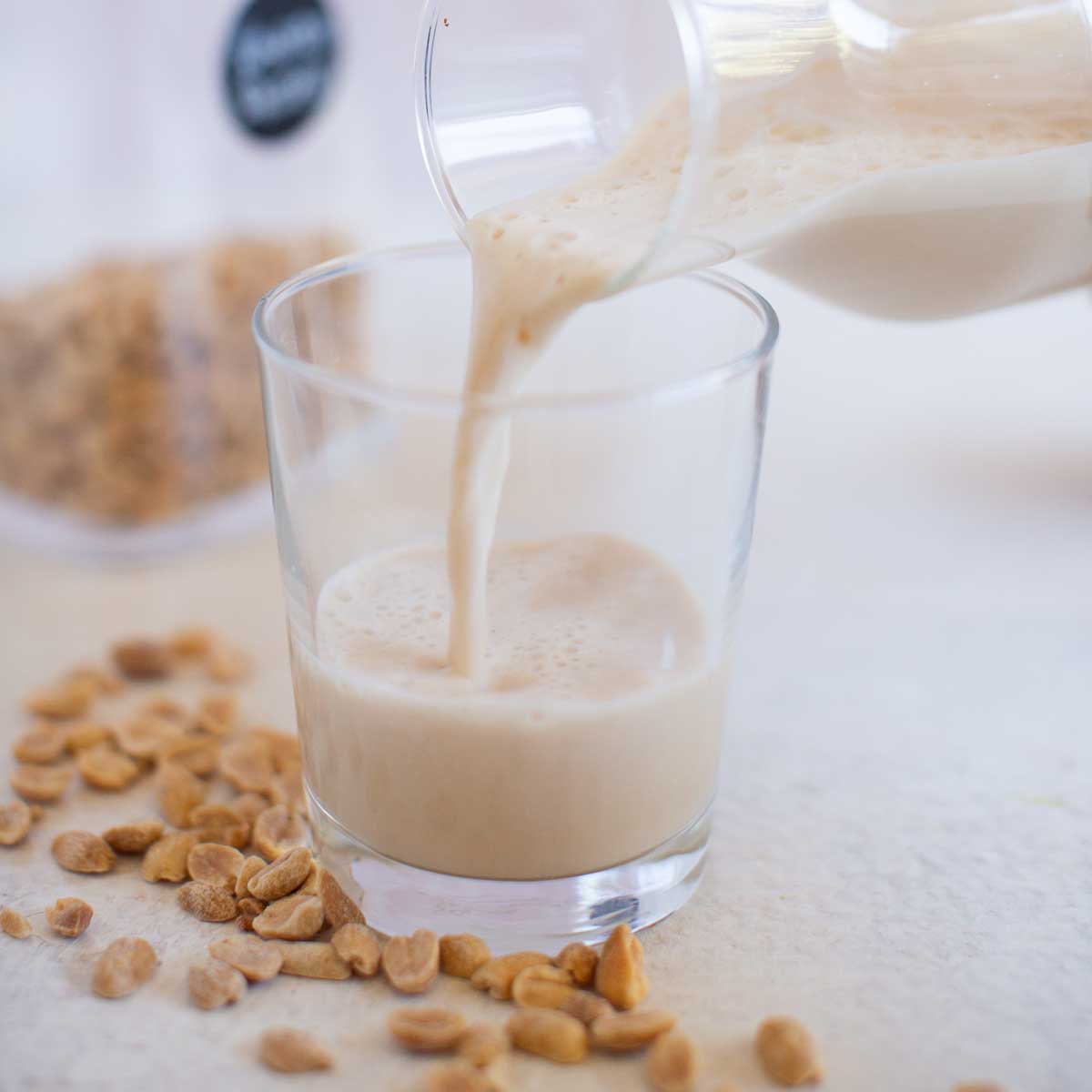 Peanut butter milk pouring from a glass bottle into a small glass with peanuts strewn around it.