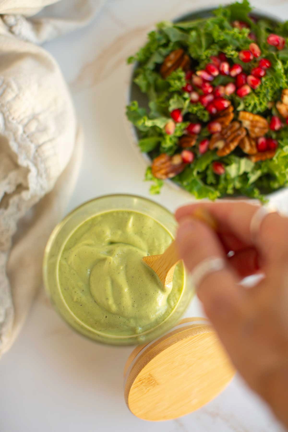 A hand stirring the final vegan avocado ranch dressing mixture in a glass jar next to a large plate of salad.