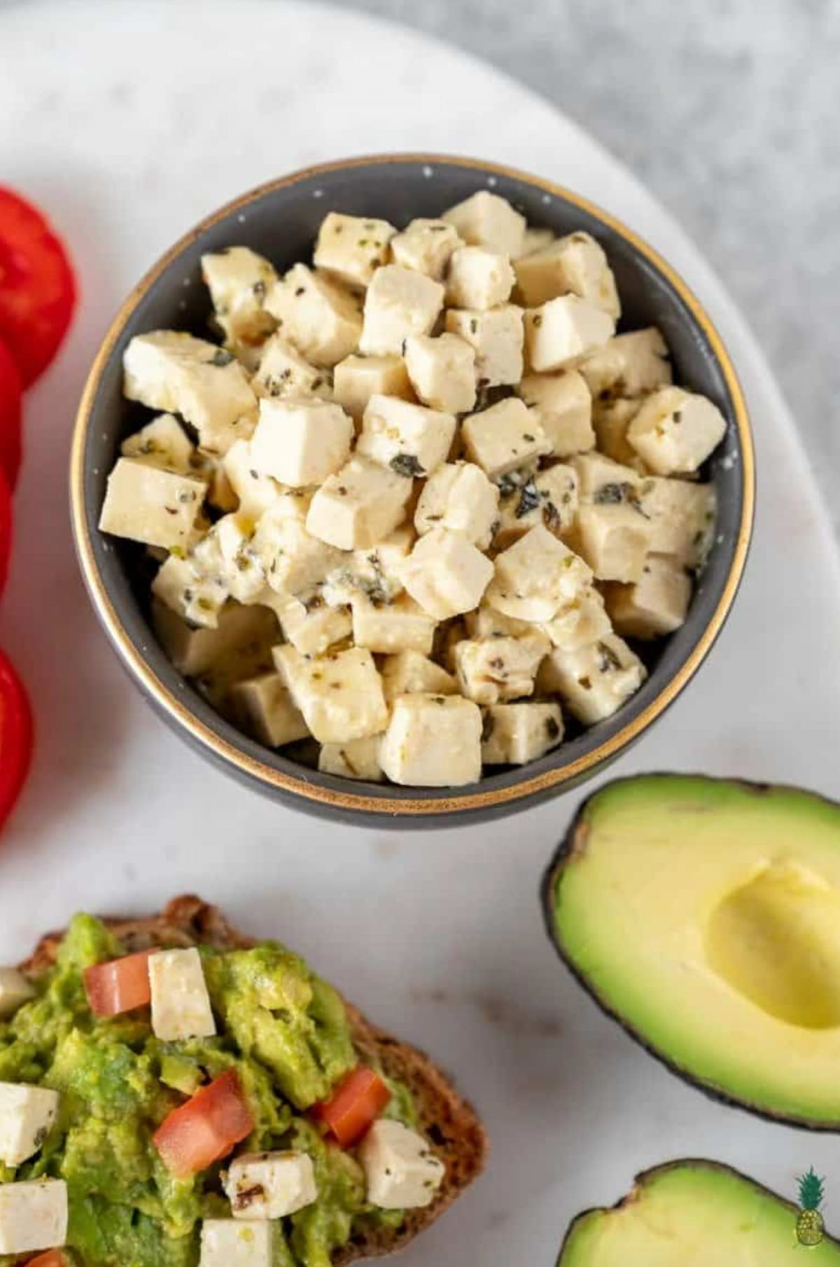 A bowl of homemade tofu feta cheese next to sliced avocados and a salad.