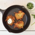 Vegan potato pancakes in a cast iron skillet next to a small bowl of fresh chopped herbs.