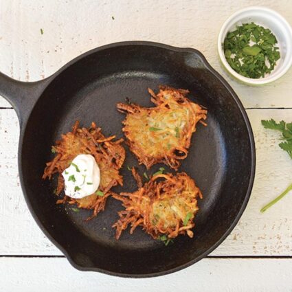 Vegan potato pancakes in a cast iron skillet next to a small bowl of fresh chopped herbs.
