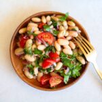 White bean salad recipe in a small wooden circular plate next to a gold fork.