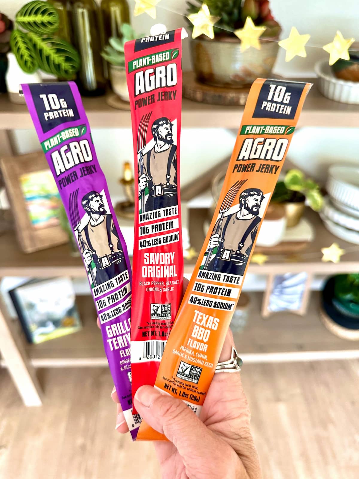 A hand holding three packages of Agro vegan jerky in flavors original, grilled teriyaki and Texas bbq.
