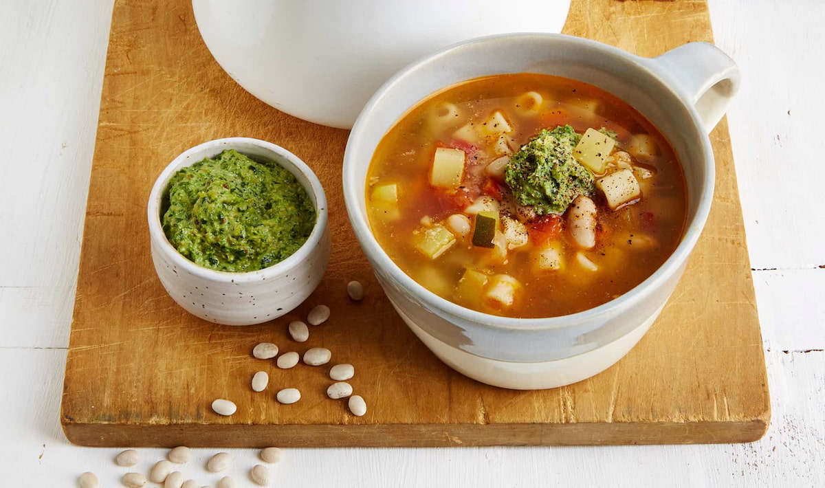 A bowl of vegan French pistou soup in a white mug and larger bowl with nuts sprinkled next to them on a cutting board.