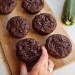 A wood cutting board with a hand reaching for chocolate zucchini muffins with a fresh zucchini next to them.