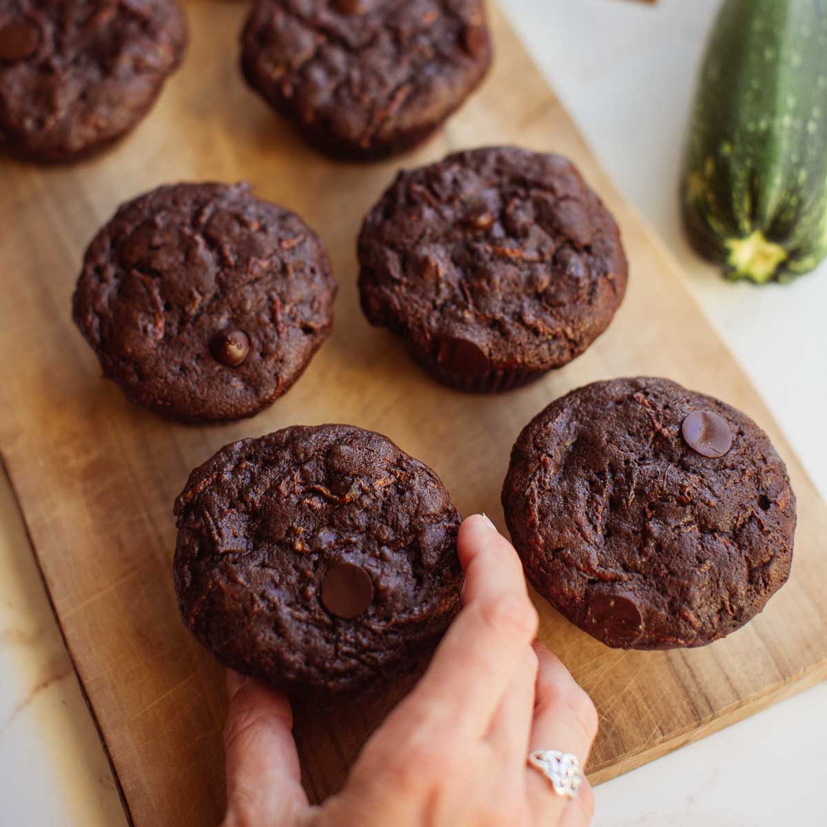 A wood cutting board with a hand reaching for chocolate zucchini muffins with a fresh zucchini next to them.