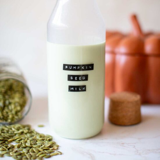 Homemade pumpkin seed milk in a bottle with black labels near a jar of pumpkin seeds laying on a countertop.