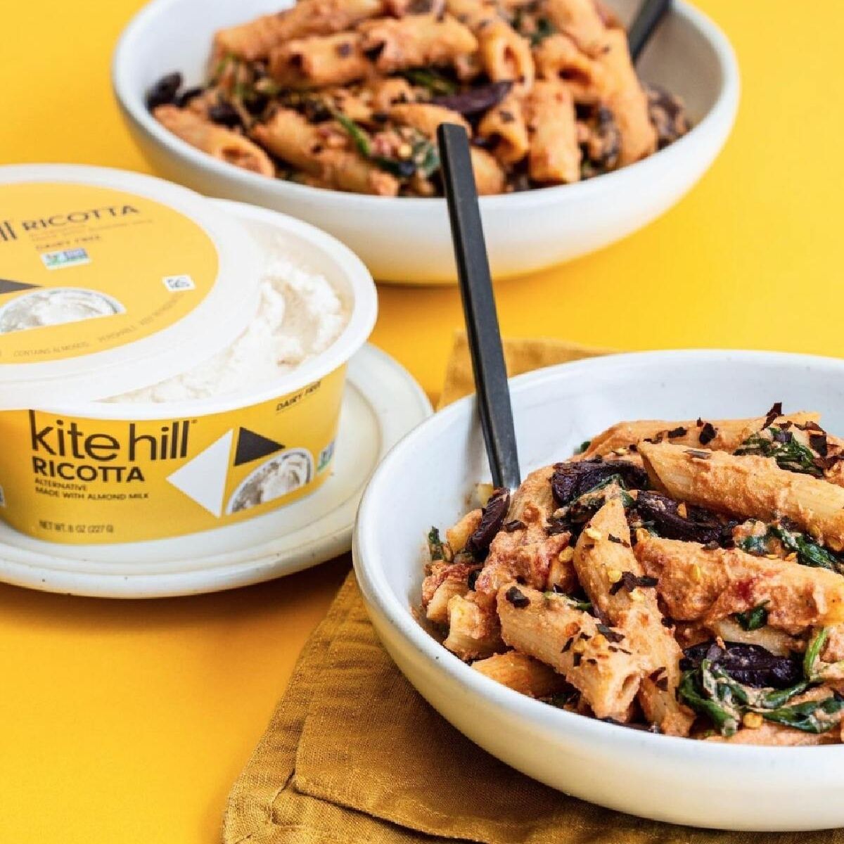 A yellow tub of Kitehill dairy-free ricotta cheese next to two bowls of pasta for a vegan ricotta cheese guide.