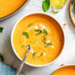 A bowl of dairy-free roasted red pepper soup with a spoon and garnished with fresh basil and soy milk.